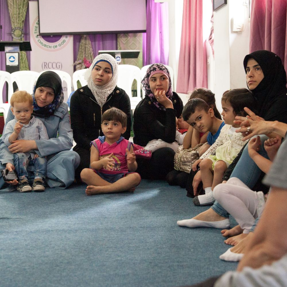 A diverse group of women and children sit on the floor in a circle, attentively listening. Various expressions and colorful attire are visible.