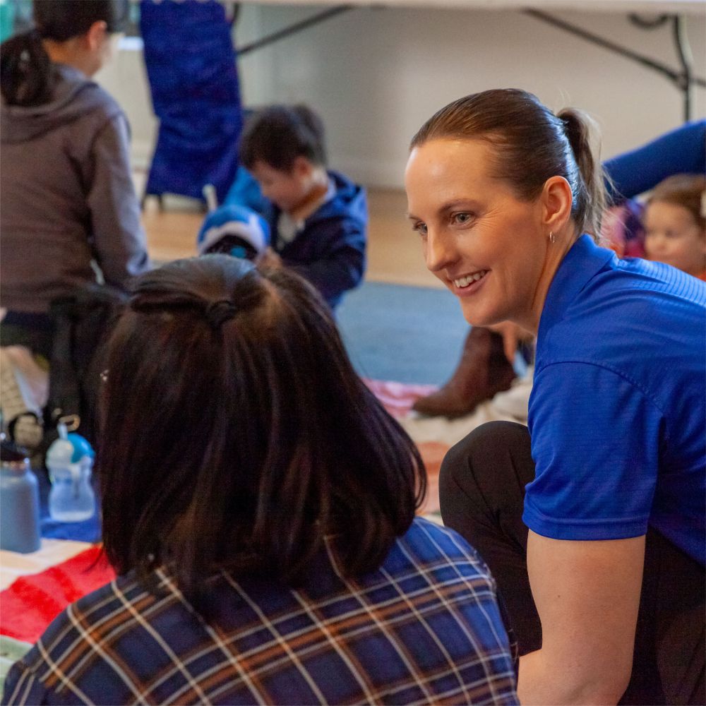 A woman in a blue shirt smiling while talking to another person; children and water bottles are in the background.