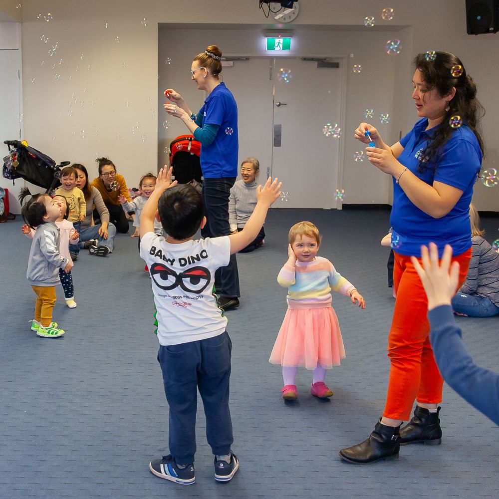 Children and adults play with bubbles in a brightly lit room. Two facilitators blow bubbles as kids reach for them, with parents watching nearby.