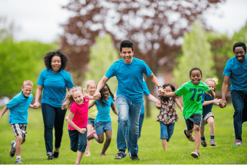 A diverse group of adults and children in bright clothing running joyfully on grass in a park, holding hands and smiling.