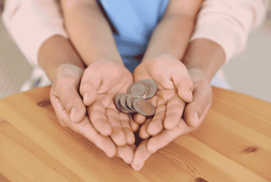Adult and child hands together holding coins over a wooden surface, symbolizing sharing or financial support.