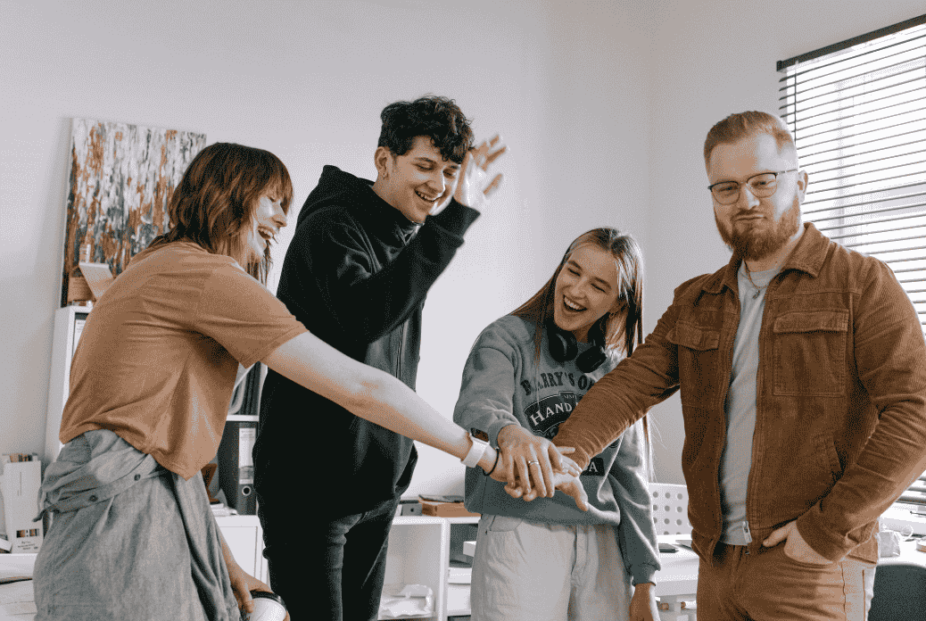 Four people smiling and joining their hands in a stack, standing in a bright room with art on the wall and blinds partially open.