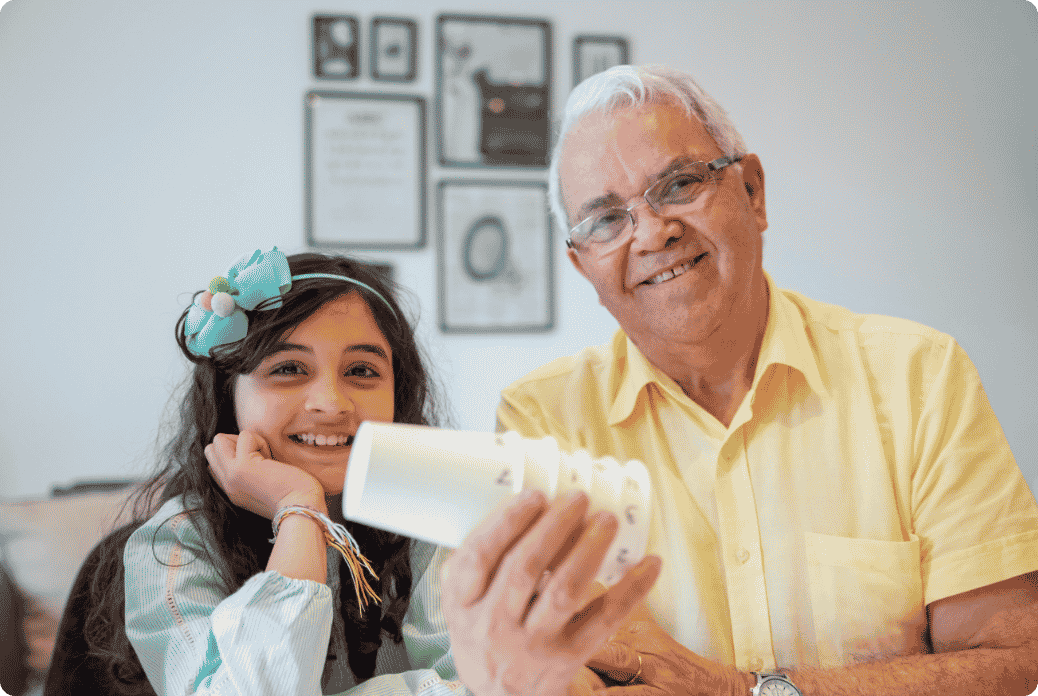 Smiling older man and young girl with a headband holding a paper object, seated in a cozy room with framed pictures on the wall.