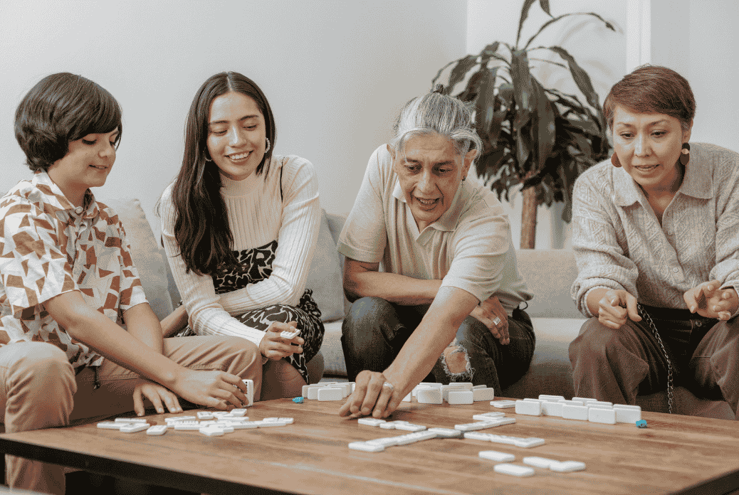 Four people sitting on a sofa playing a game of dominoes on a wooden table, smiling and engaged in the activity.
