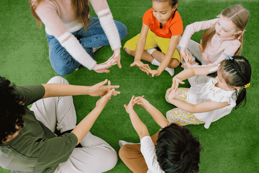 Children and two adults sit in a circle on green grass-like carpet, playing a clapping game with focused expressions.