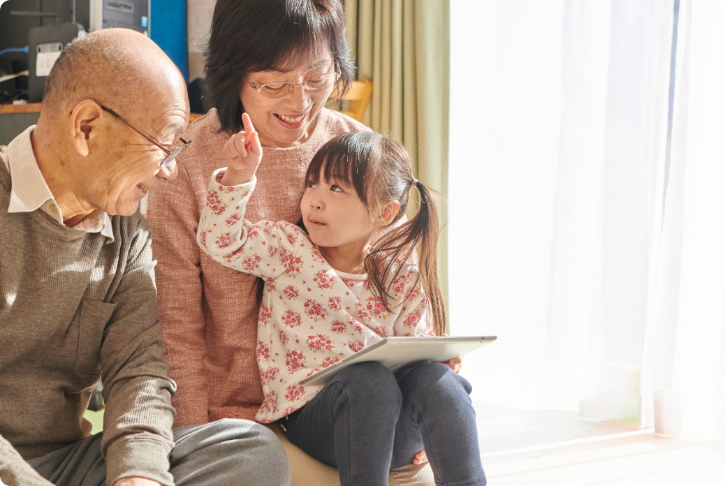 A young girl holding a tablet sits on the lap of a smiling woman, while they talk with an elderly man indoors, by a sunlit window.