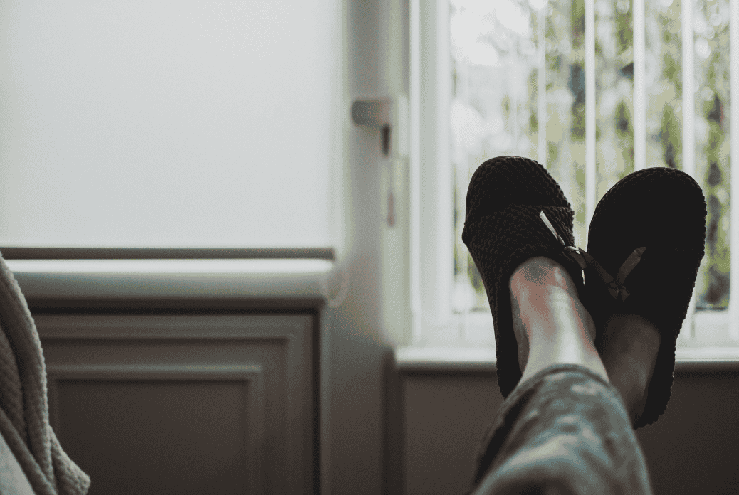 Person relaxing with feet up, wearing cozy slippers, near a bright window with vertical blinds partially open.