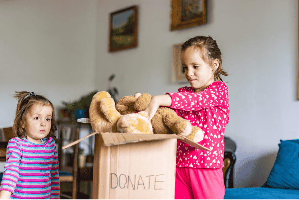 Two young children packing a cardboard box labeled "DONATE" with stuffed animals in a cozy living room.