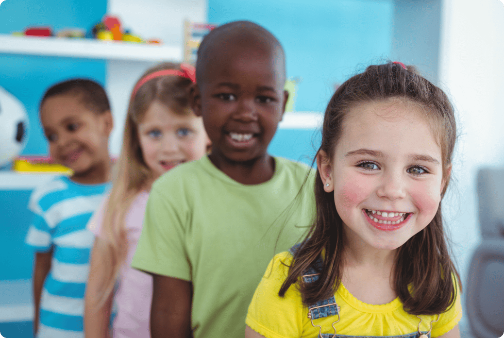 Four smiling children stand in a line indoors, with a shelf and toys in the background.