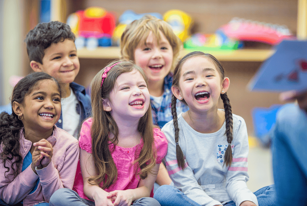 Five children sitting and laughing together in a classroom, looking towards a person holding a book, with colorful toys in the background.