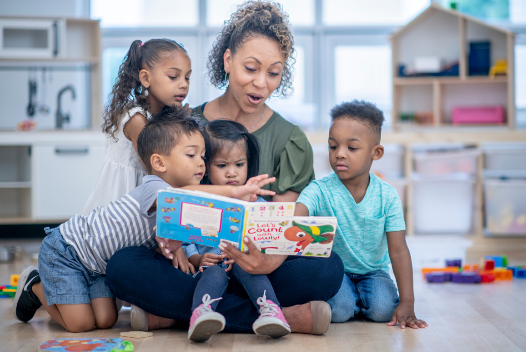 A woman reads a colorful book to four young children in a bright classroom filled with play furniture and toys.