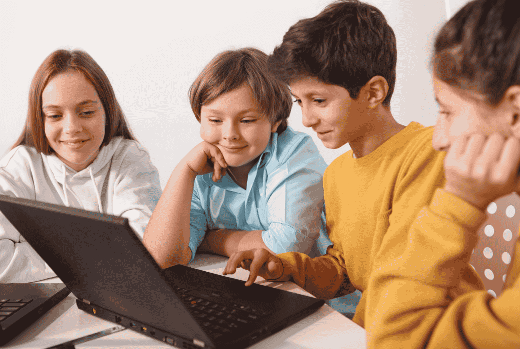 Four children are gathered around a laptop, smiling and engaged. They appear to be focused on something on the screen.