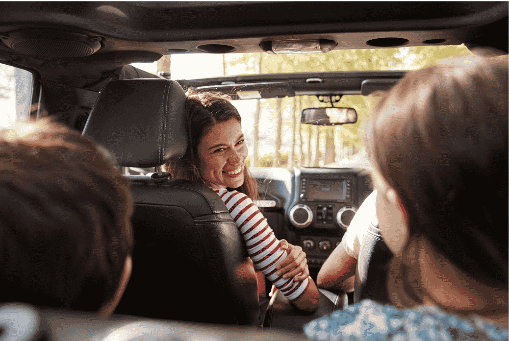 Smiling woman in the driver's seat of a car, looking back at two kid passengers under a sunlit, tree-lined road.