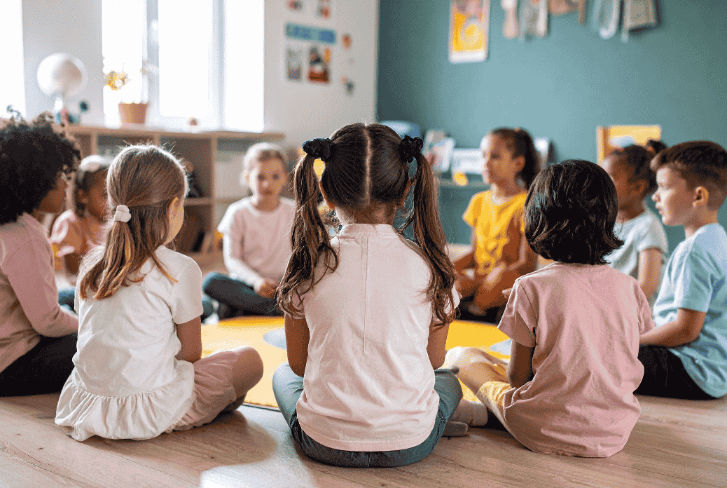 Children sitting in a circle on a classroom floor, engaged in a group activity, with colorful walls and shelves in the background.