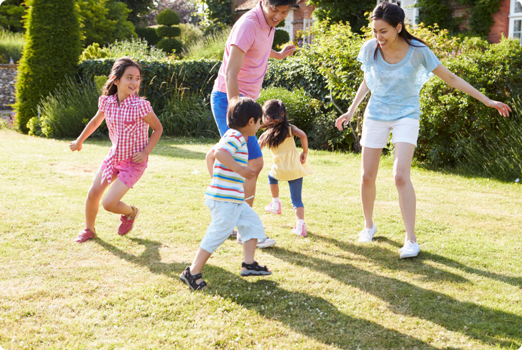 A family plays tag on a sunny day in a garden, with children running and adults joyfully joining in.