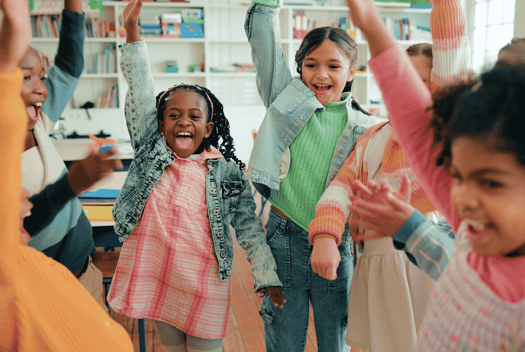 Children in a classroom joyfully raise their hands, smiling and celebrating together. Shelves with books are visible in the background.