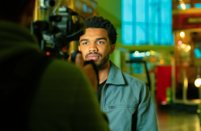 A person being filmed by a camera indoors, with blurred colorful lights in the background.