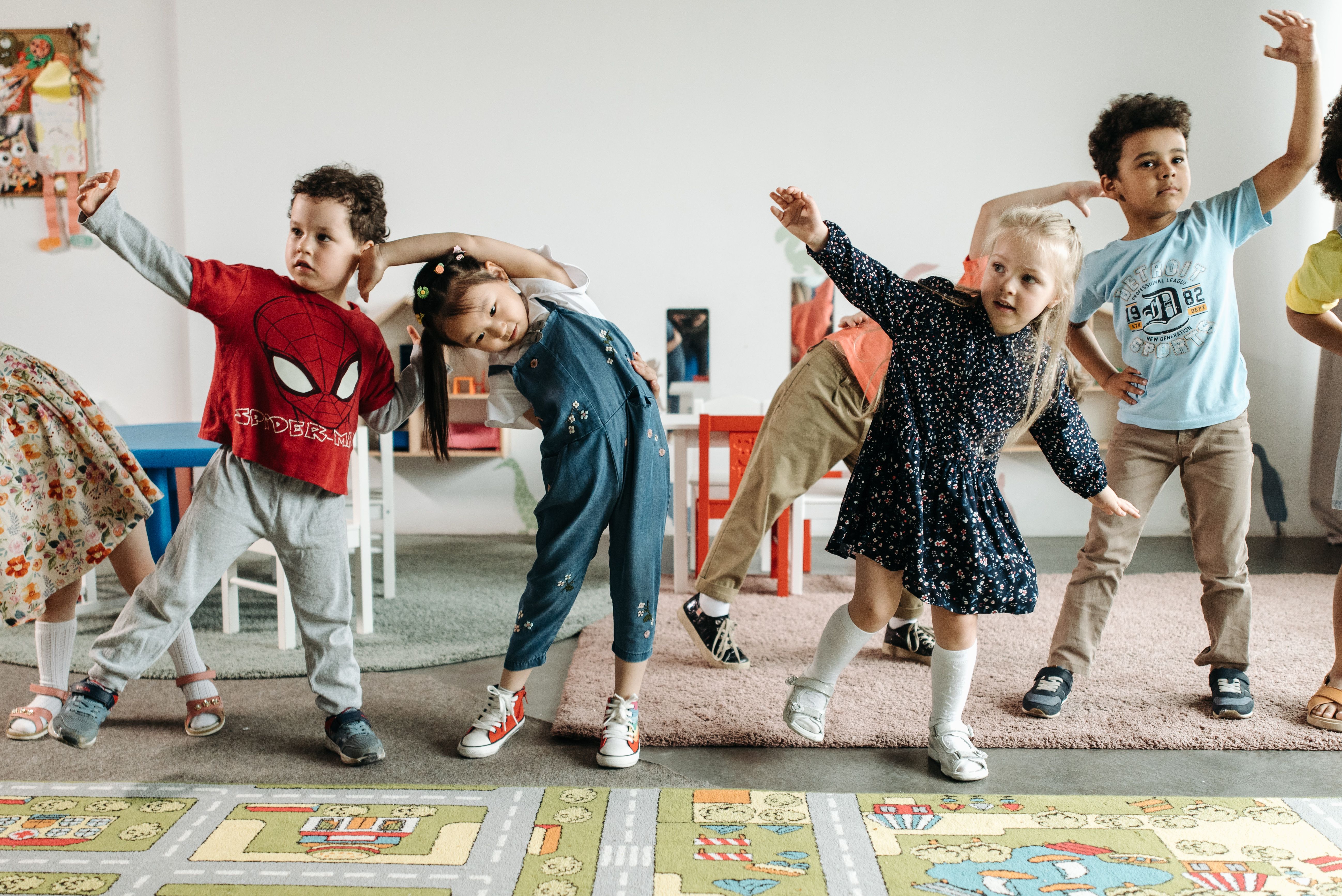 Children are playfully stretching and posing in a classroom, with colorful rugs and furniture in the background.