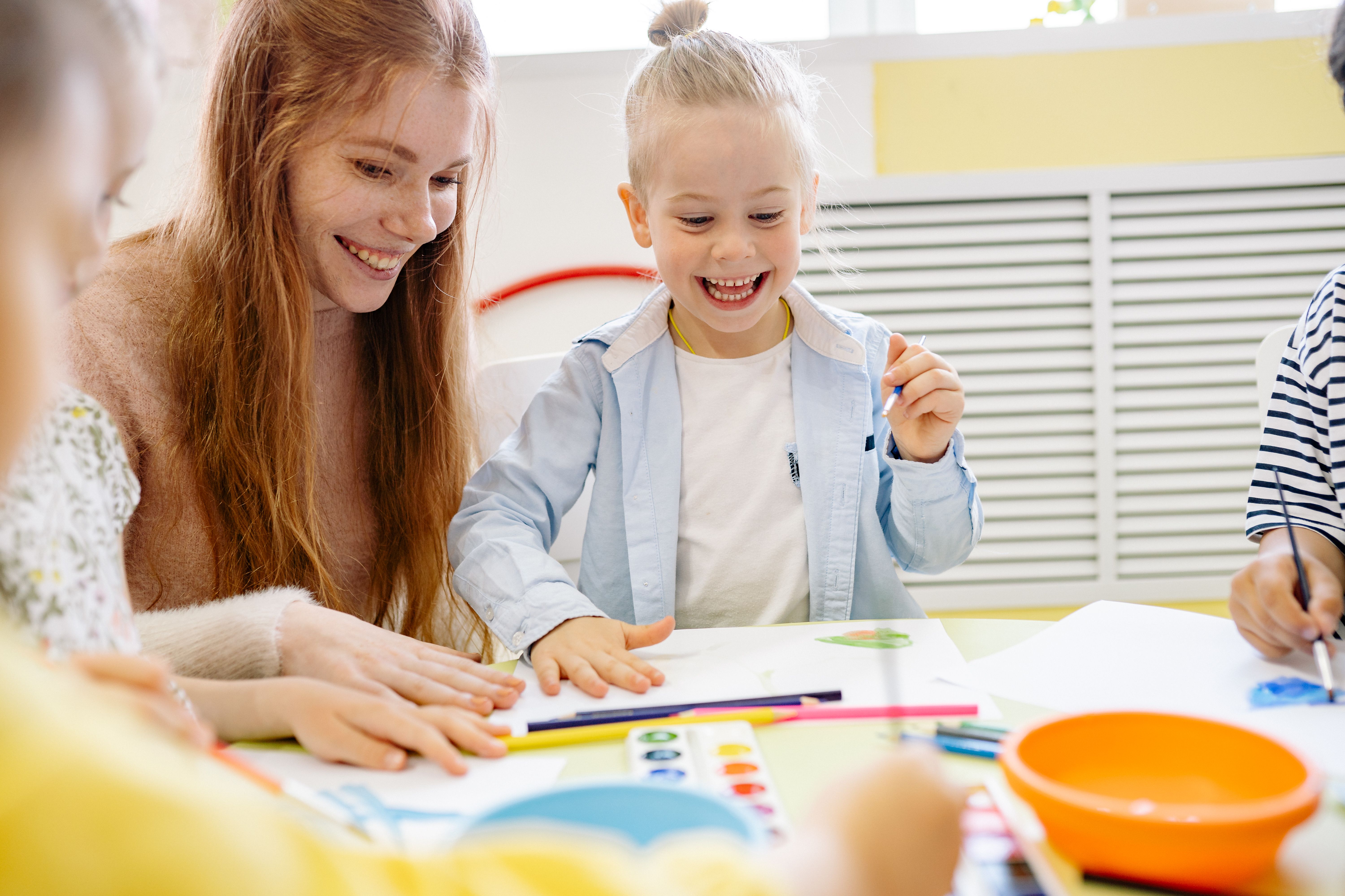 A woman and children smiling while drawing and painting at a table with colored pencils and paints in a bright room.
