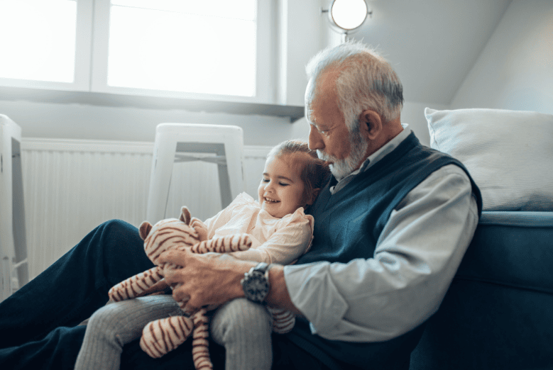 Elderly man and young girl sitting on the floor, smiling and holding a stuffed animal in a cozy, sunlit room.