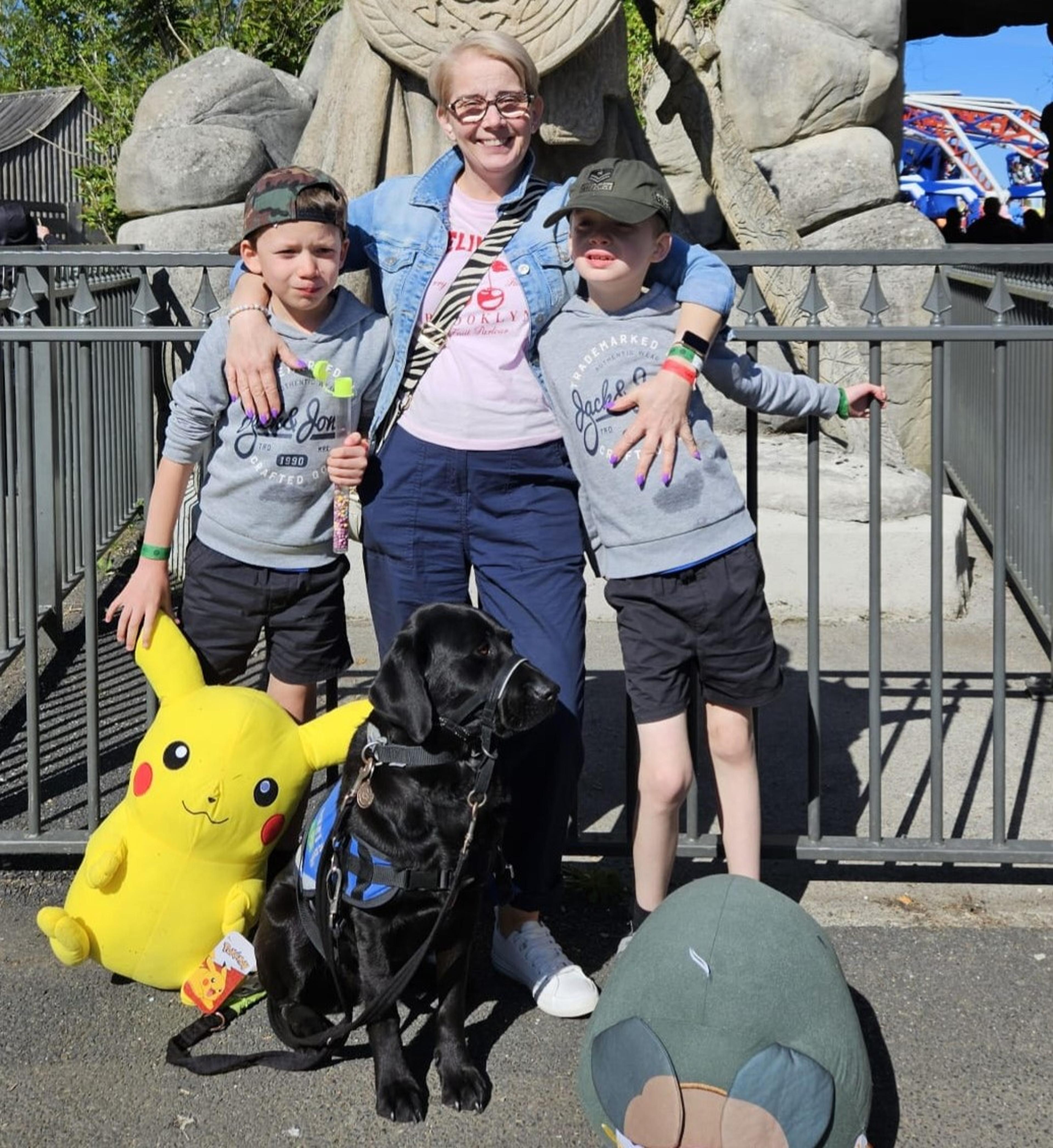 Sarah standing in a park with arms around both of her sons and Kendall sitting on the ground in front. One child is holding a large Pikachu teddy
