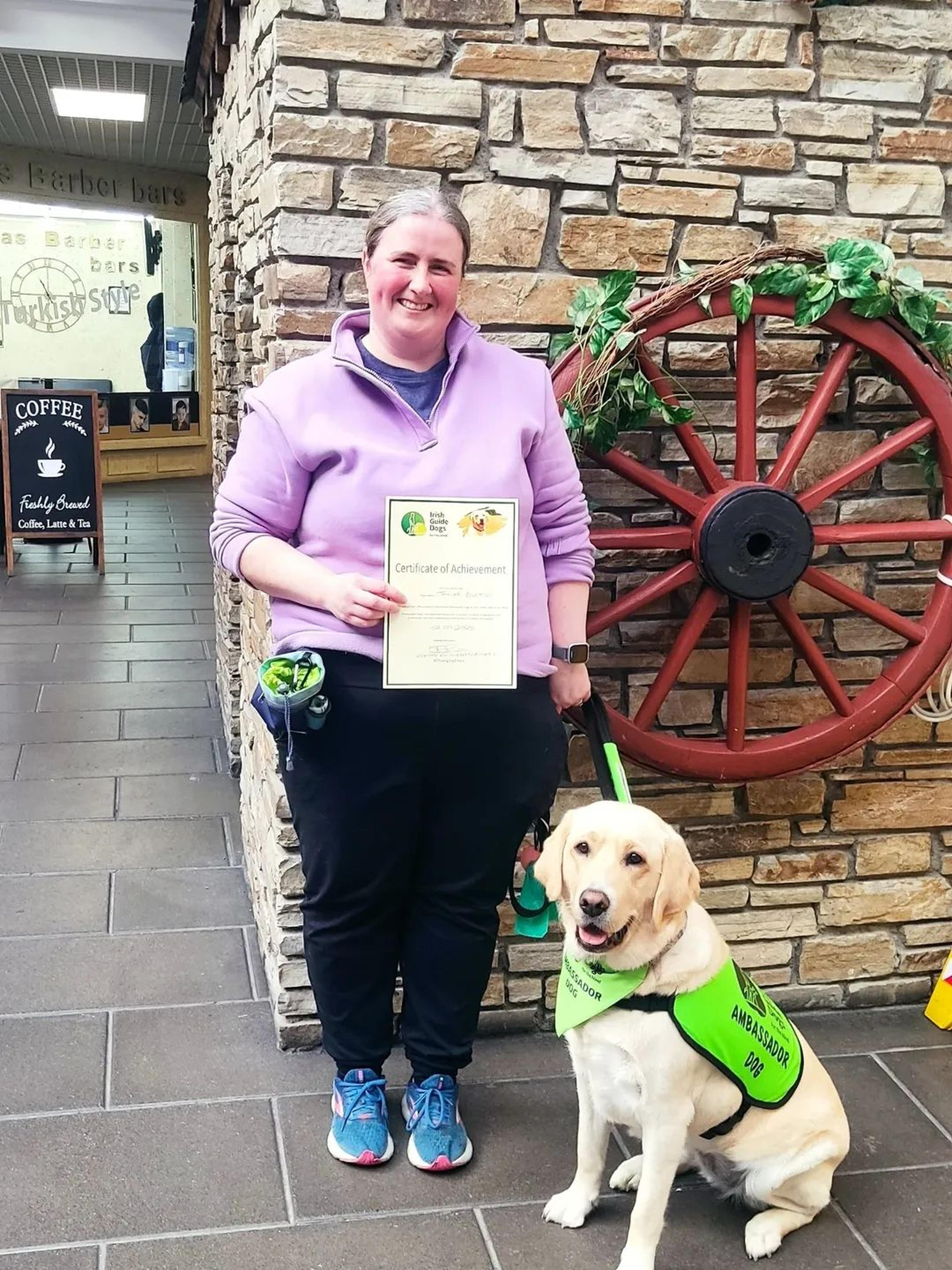 Rachel standing holding Ambassador Dog certificate, with Taylor sitting at the side