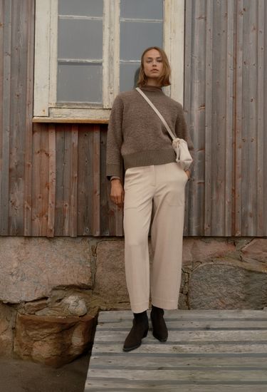 Woman in a brown sweater and beige pants stands on wooden steps against a rustic wooden wall, with a crossbody bag and black boots.