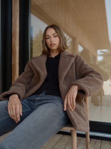 Person sitting on a wooden chair, wearing a brown coat and jeans, with a glass building in the background.