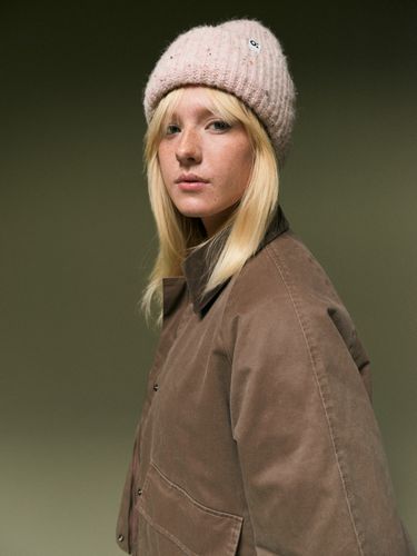 Person with long blonde hair wearing a pink knit hat and brown jacket, looking to the side against a neutral background.