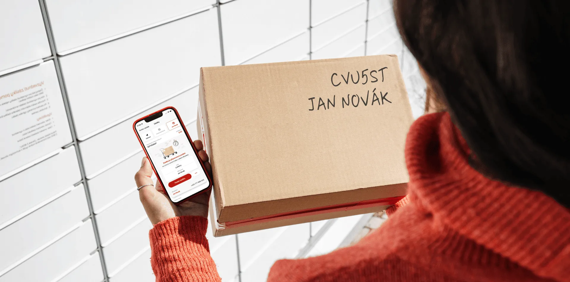 Osoba držící chytrý telefon s doručovací aplikací a kartonovou krabici označenou jménem, stojící před zdí.