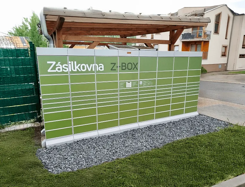 Zelený parcel locker s označením „Zásilkovna Z-BOX“ na štěrkovém podloží, poblíž dřevěného přístřešku a obytných budov.