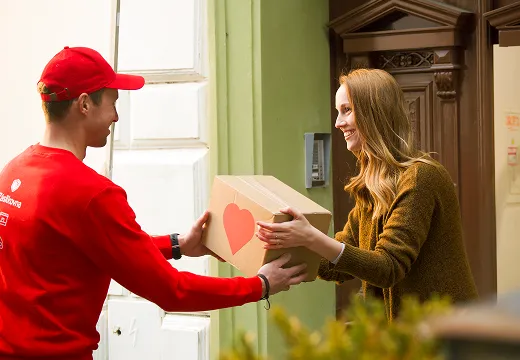 A delivery person in a red uniform hands a cardboard box with a heart design to a smiling woman at her doorway.