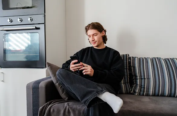 A person in a black sweater sits on a couch, looking at a smartphone, near a modern kitchen oven.