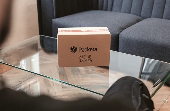 A cardboard package labeled "Packeta" and addressed to "Jan Novak" sits on a glass coffee table with a couch in the background.