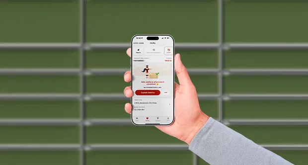 Ruka drží chytrý telefon se zobrazenou aplikací pro sledování zásilky. Na pozadí je rozmazaná zelená stěna s vodorovnými liniemi.