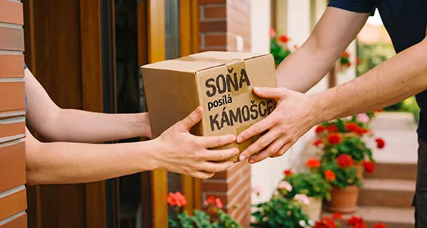 Two people exchanging a package labeled "SOŇA posílá KÁMOŠCE" in front of a brick house with flowers.