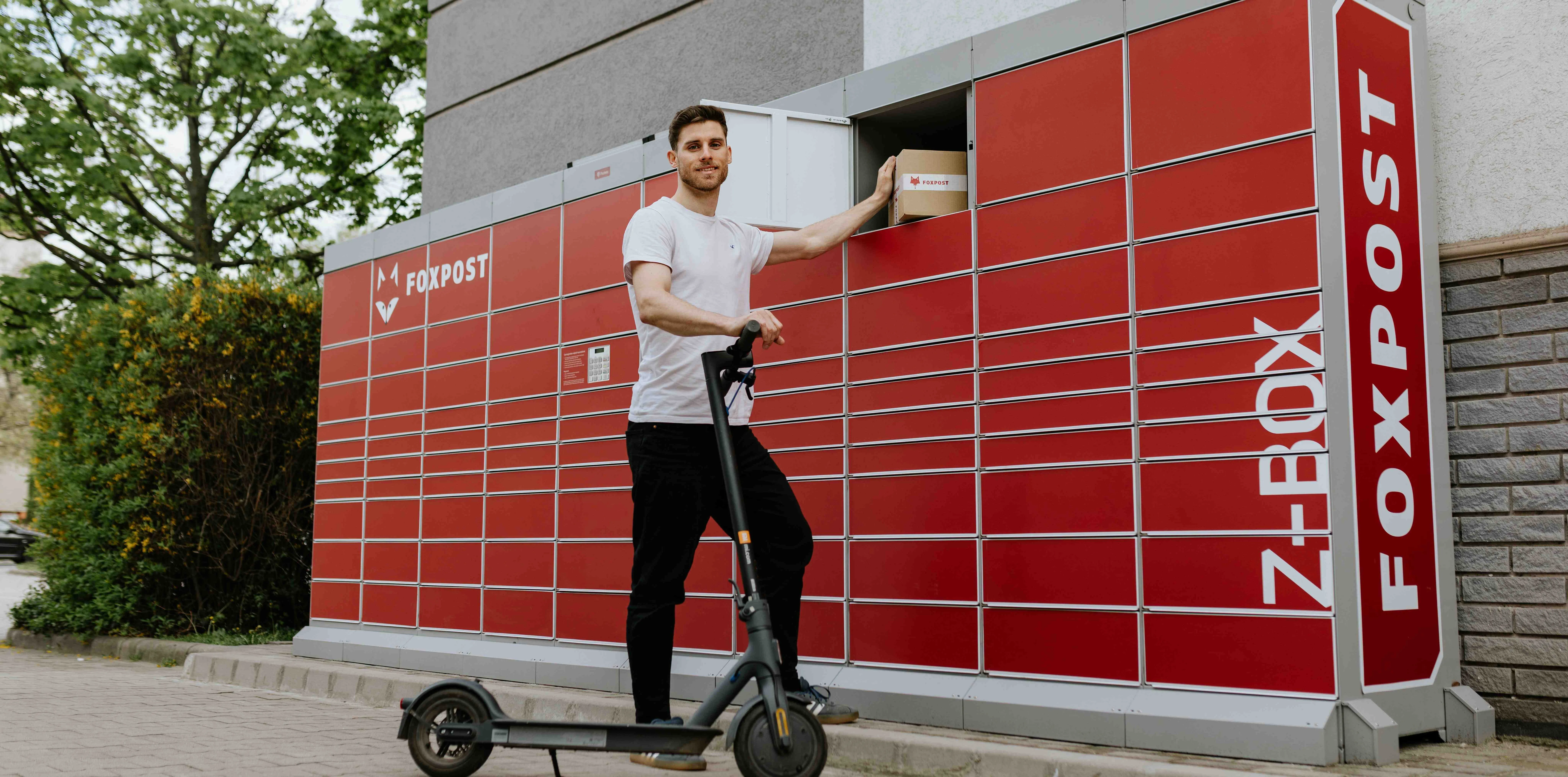 Man retrieving a package from a red Foxpost locker while standing with an electric scooter.