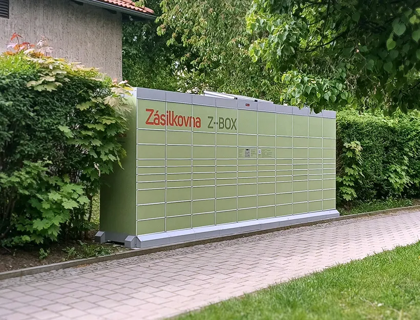 Výdejní box Zásilkovna Z-BOX umístěný venku vedle domu, obklopený zelení a situovaný podél dlážděné cesty.
