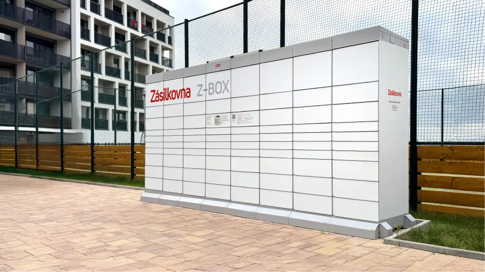 Venkovní parcelní skříňka s označením „Zásilkovna Z-BOX“ u moderní budovy, se zpevněným chodníkem a oploceným areálem.