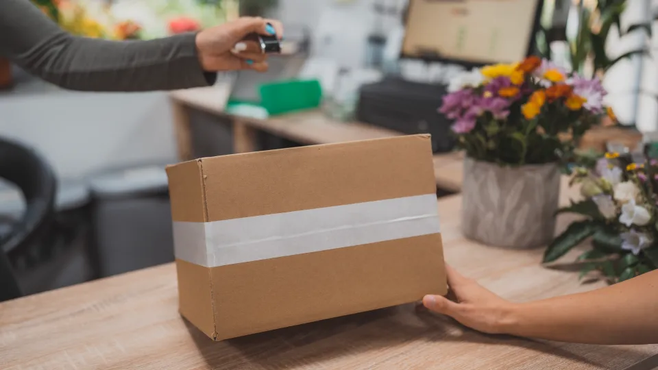 A package is being scanned by a hand-held device over a counter with flowers in the background.