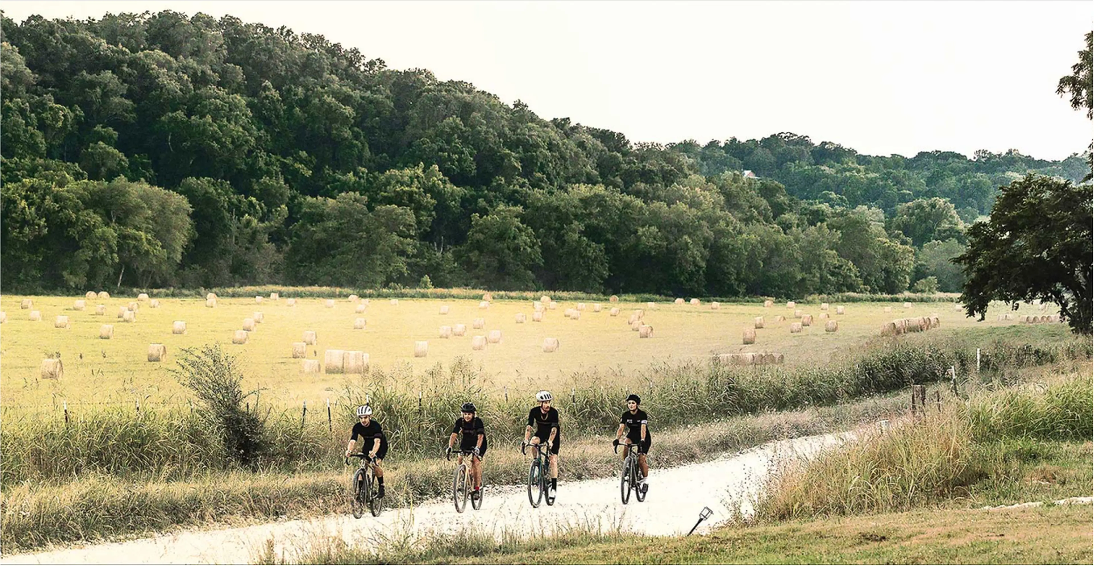 Four cyclists ride on a gravel path through a scenic rural landscape with hay bales and lush green trees in the background.
