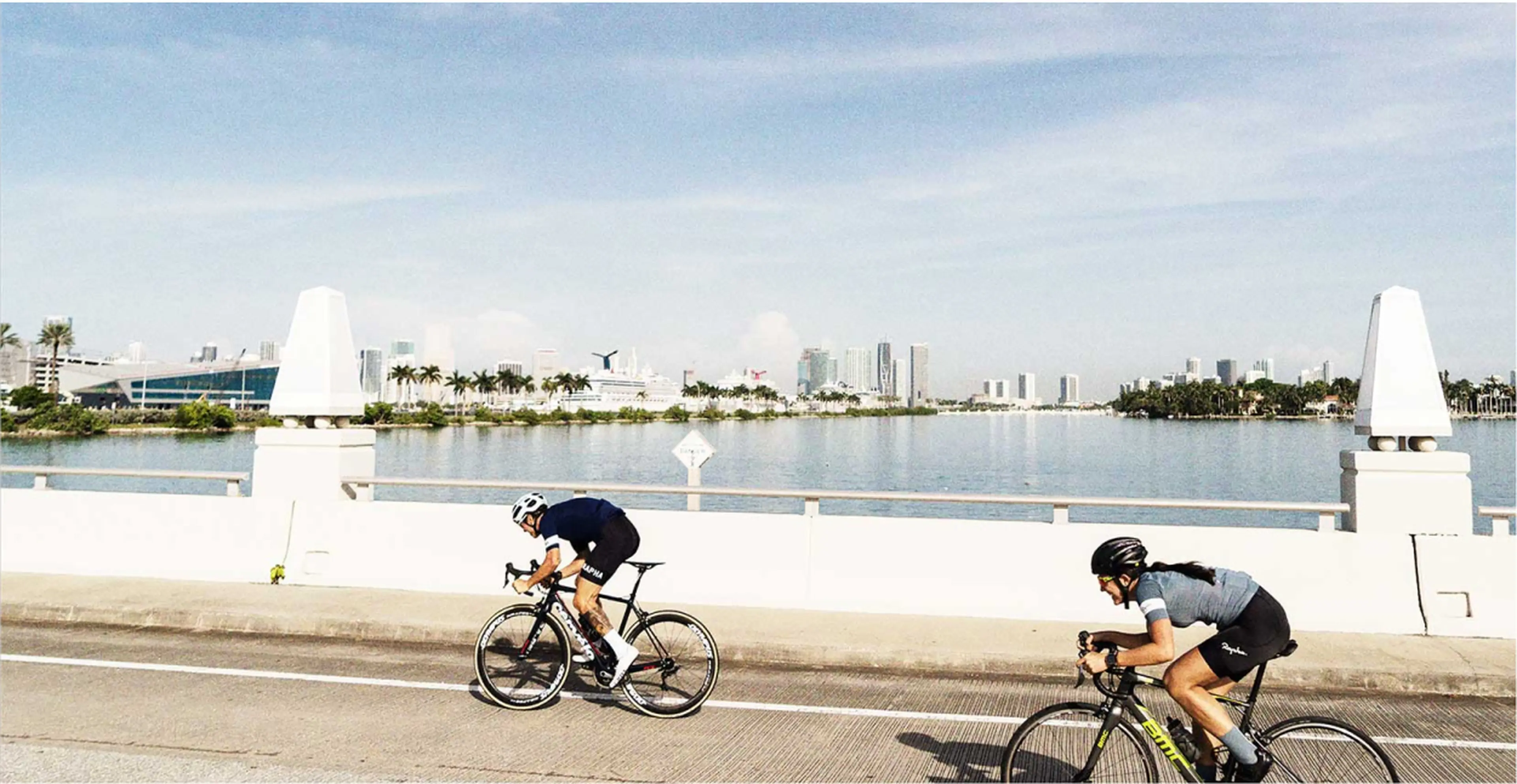Two cyclists ride on a bridge with a city skyline and water in the background under a clear blue sky.