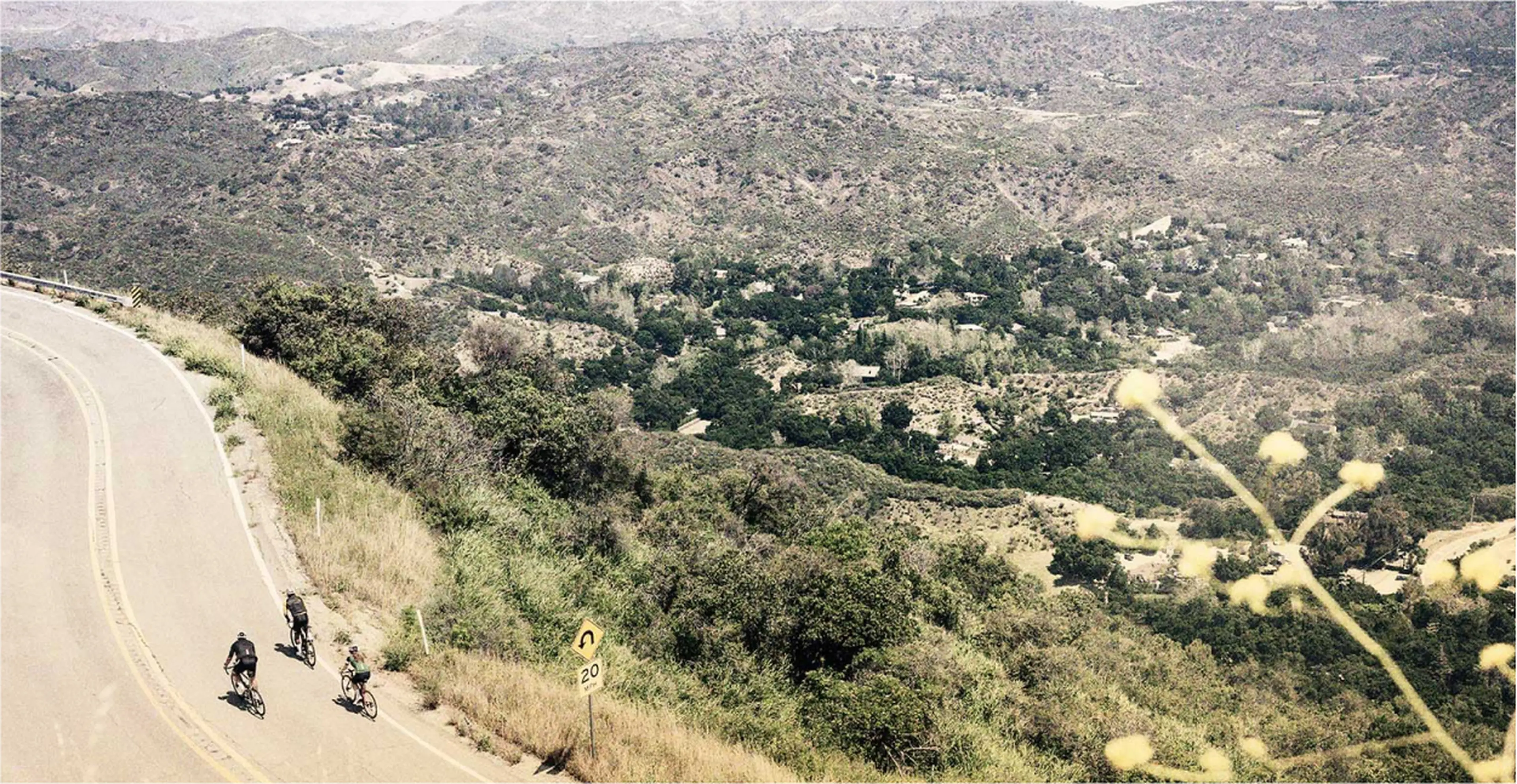 Three cyclists ride along a winding mountain road, surrounded by lush greenery and distant hills under a hazy sky.