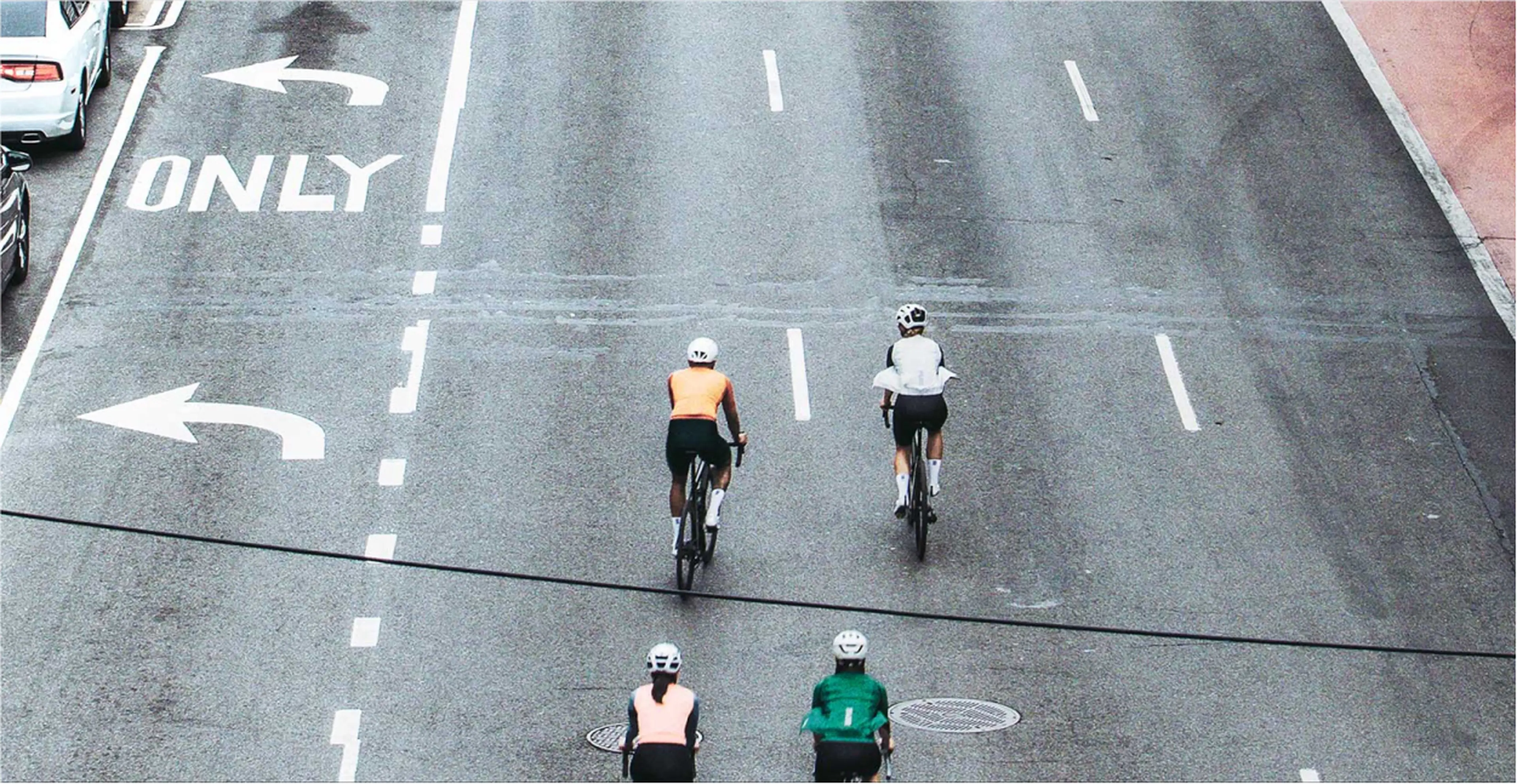 Four cyclists ride on a city street with marked lanes, including a left-turn-only lane. Cars are parked on the left side.