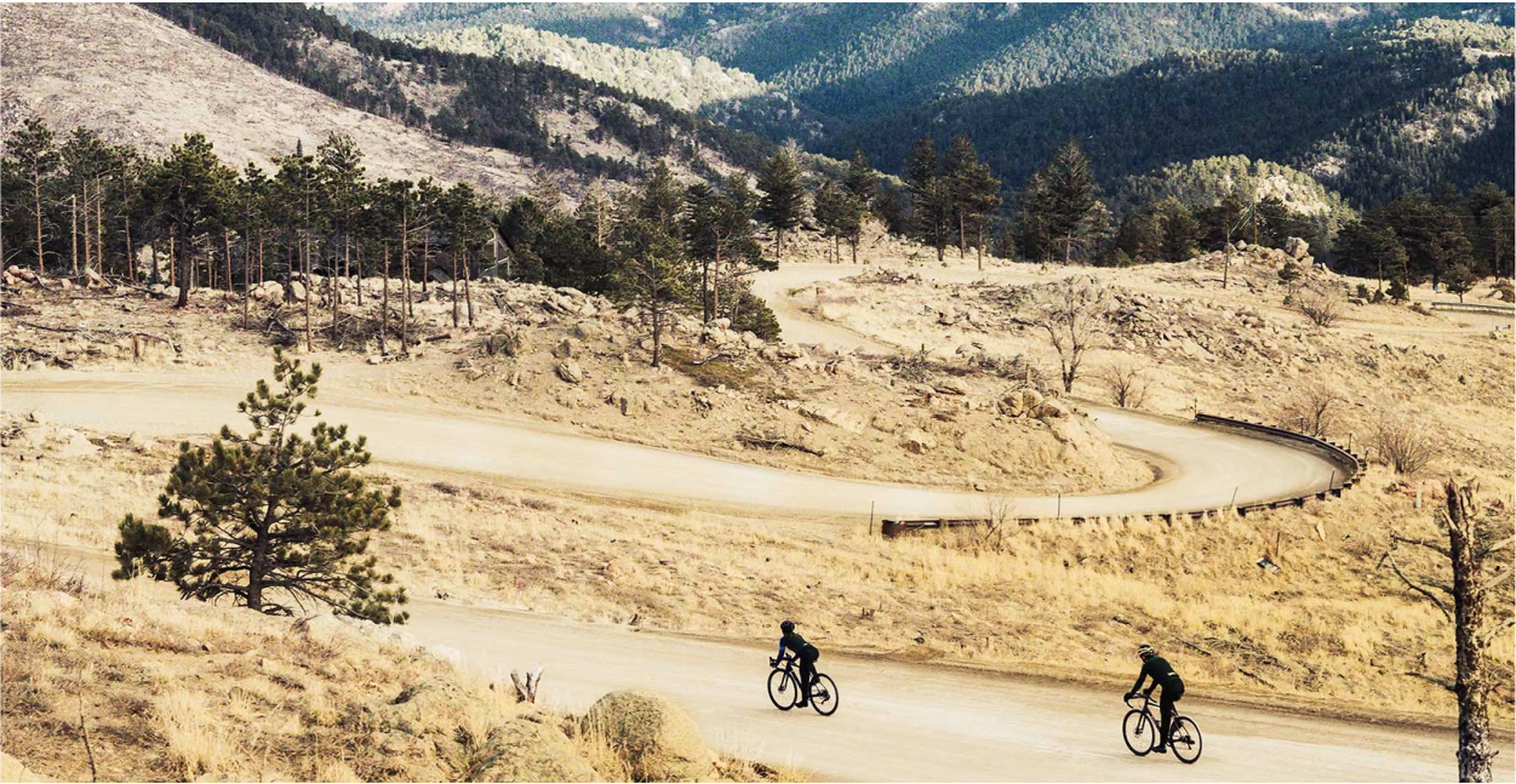 Two cyclists ride on a winding mountain road surrounded by rocky terrain and sparse trees under a clear sky.