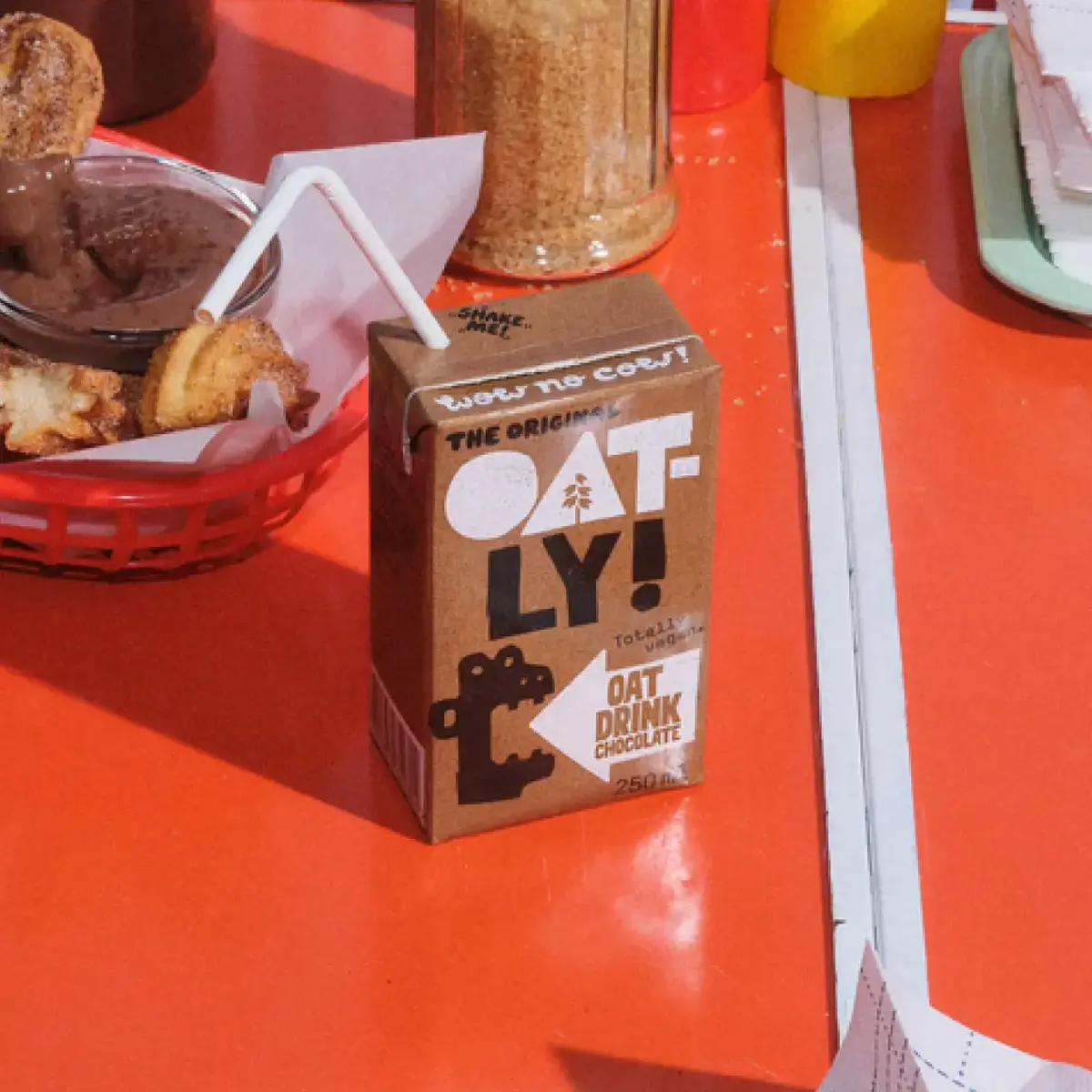 Carton of Oatly chocolate oat drink with a straw on a red table, surrounded by snacks and condiments.