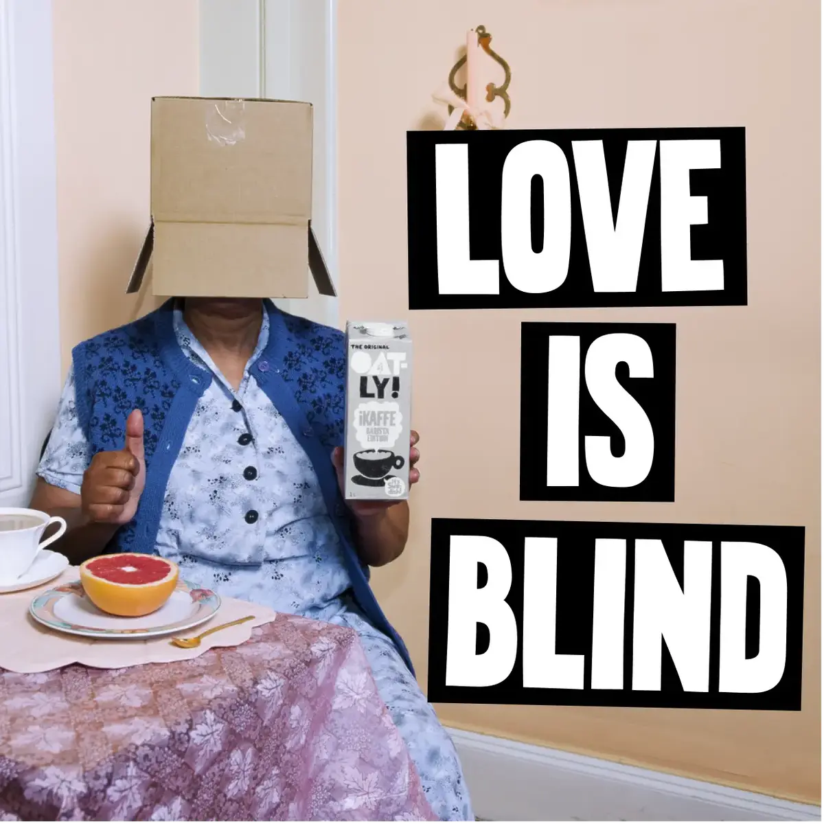 Person with a box on their head gives a thumbs up, holding oat milk. Text reads "LOVE IS BLIND." Grapefruit and tea on the table.