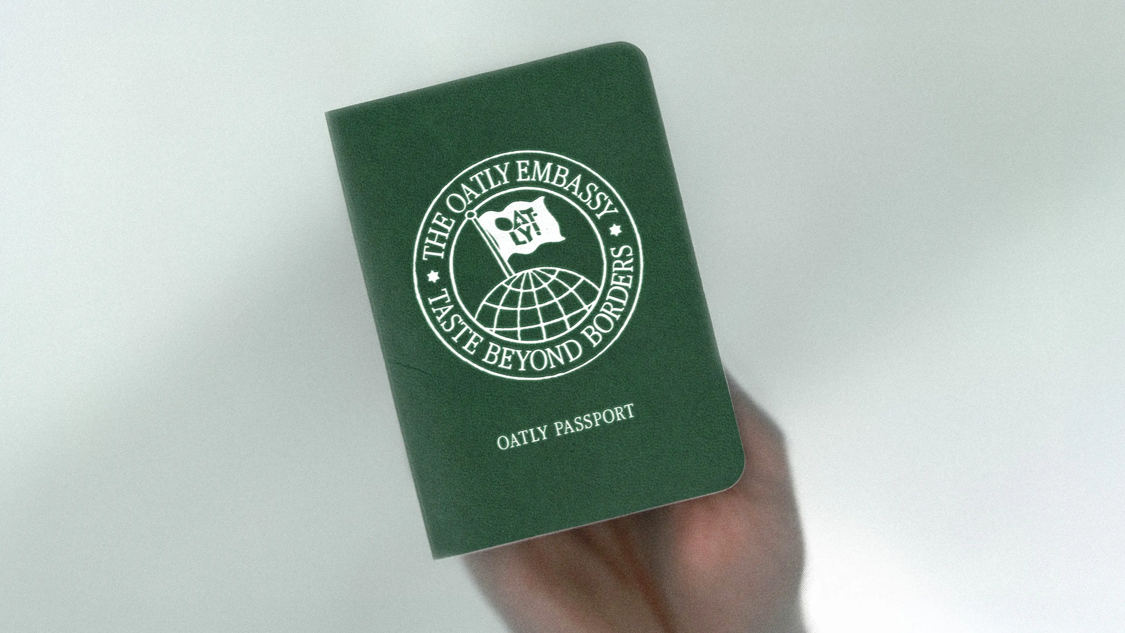 A hand holds a green booklet labeled "Oatly Passport" and "The Oatly Embassy Taste Beyond Borders" with a globe and flag illustration.