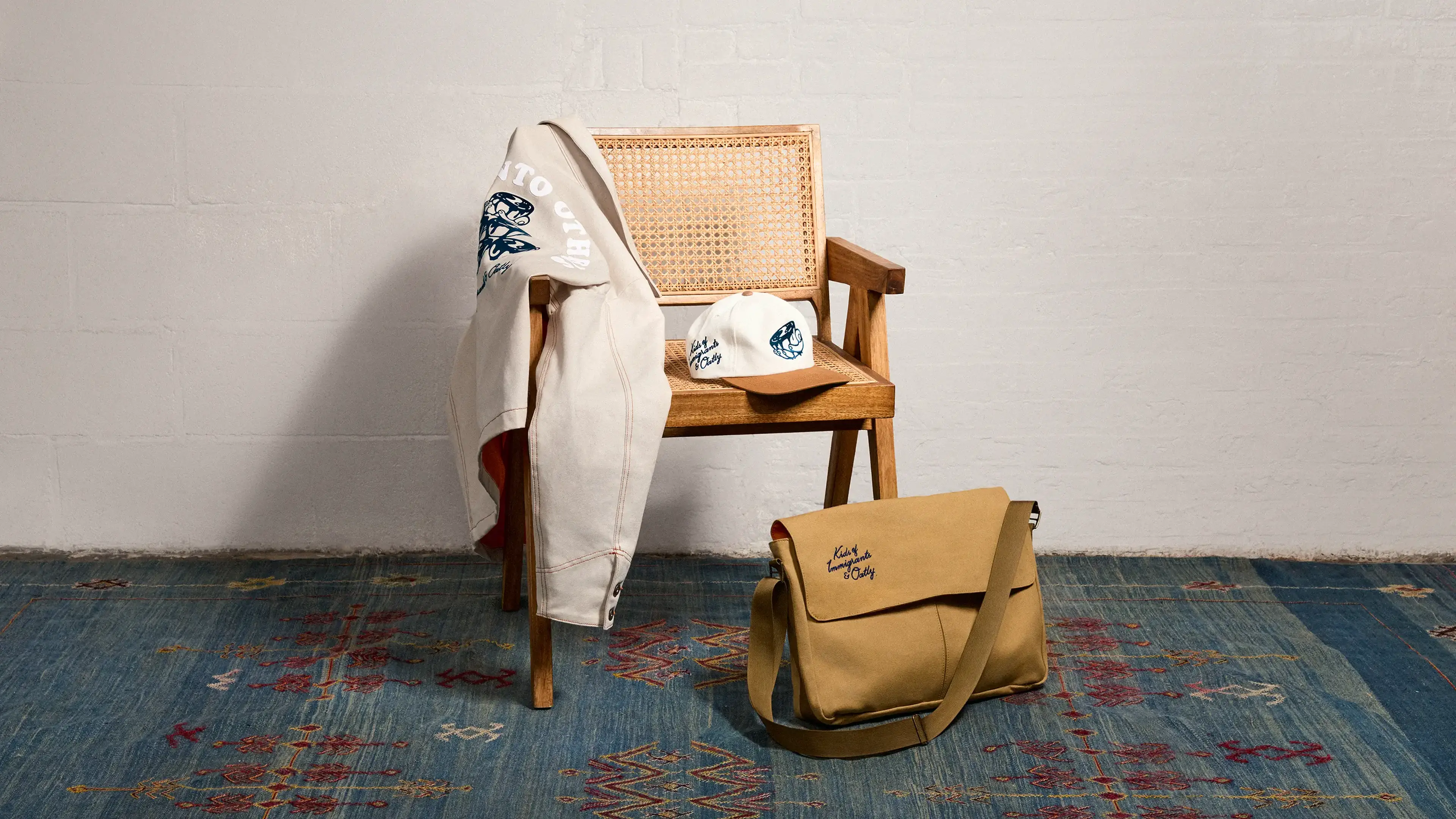 Wooden chair with beige jacket and cap draped over it, and a tan messenger bag on the floor, against a white wall on a patterned blue rug.