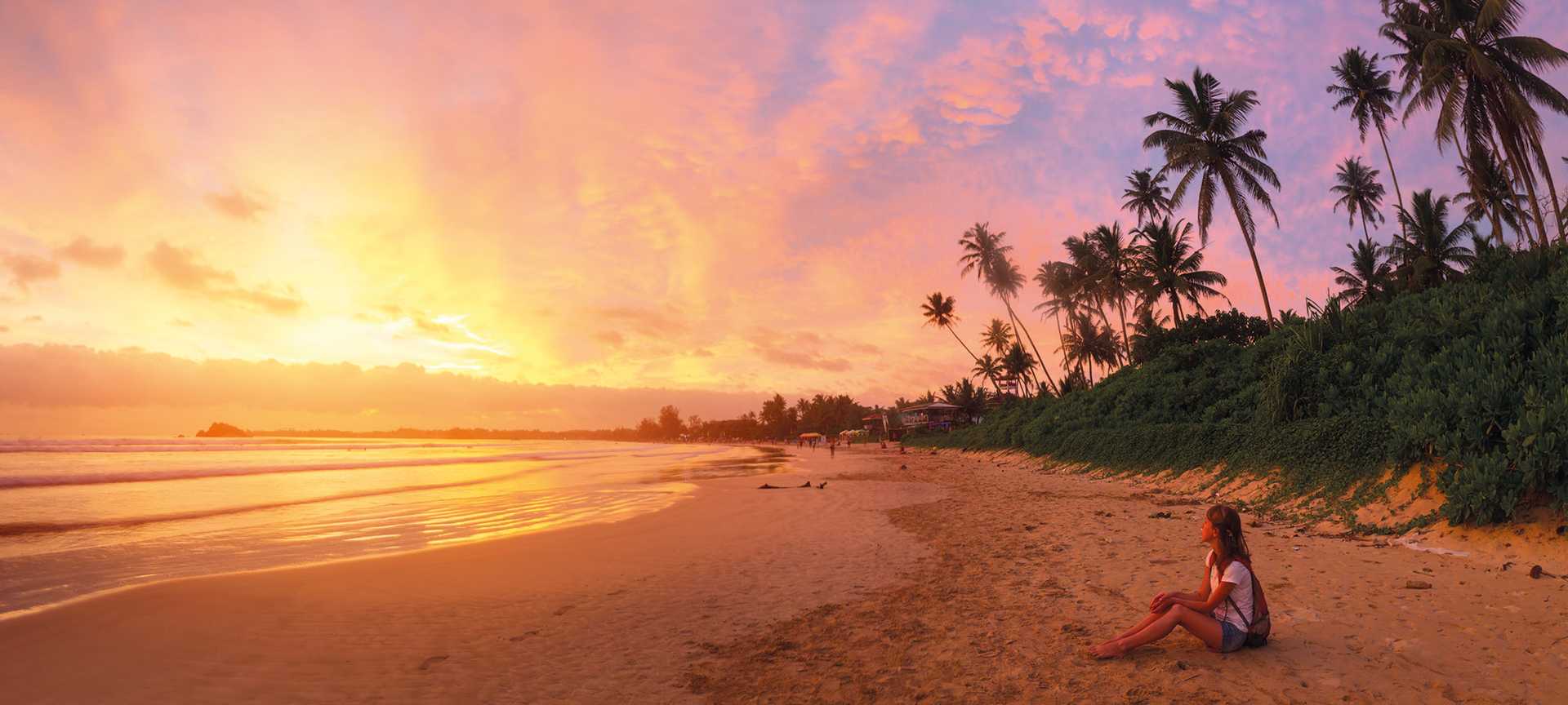 A woman sits on a sandy, palm-lined beach in Sri Lanka to watch the sunset